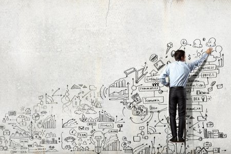 Businessman on Ladder Drawing Business Concepts on Wall